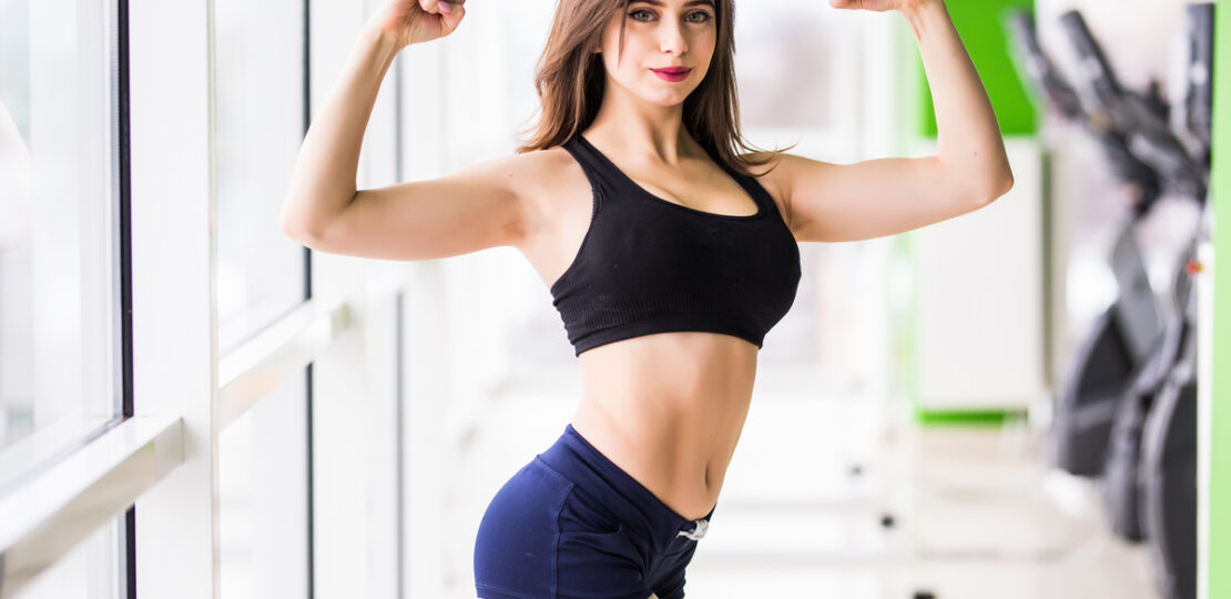 beauty with big green eyes long brown hair is posing in gym in front of the windows